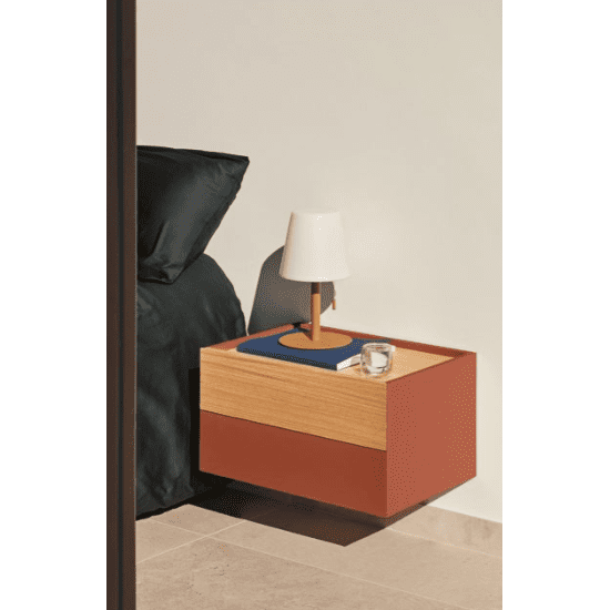 La mesita de noche Otto 2C (Suspendida), con un diseño en madera y rojo, permite guardar de forma elegante una pequeña lámpara, un libro y un vaso de agua. Se ubica perfectamente junto a una cama con ropa de cama oscura.
