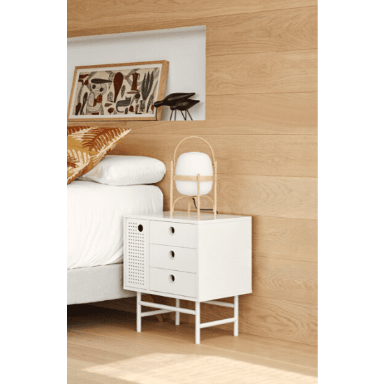 La Mesita de noche blanca Punto 1P3C, con tres cajones, una lámpara moderna y decoración en la parte superior, ofrece un diseño contemporáneo que complementa los paneles de madera de la pared y la cama con sábanas blancas del fondo.
