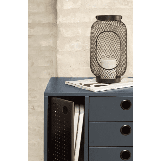 Sobre una mesita de noche azul Punto 1P3C, que cuenta con tres cajones y almacenamiento interno que contiene un cuaderno junto a dos revistas, se encuentra una vela dentro de un soporte de metal negro, resaltando el diseño contemporáneo.