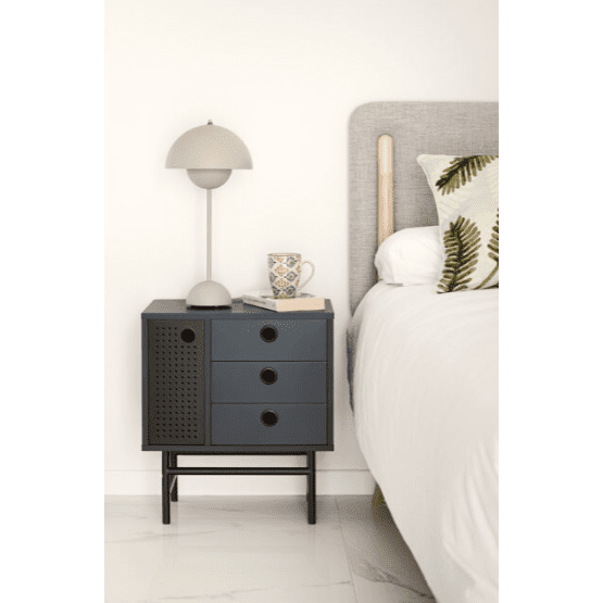 Escena de dormitorio moderno con una mesita de noche Punto 1P3C pequeña y oscura con tres cajones y una lámpara. Sobre ella hay una taza de café. La cama tiene un cabecero beige y una almohada con estampado de helechos, que realzan el diseño contemporáneo con suelo de baldosas blancas.