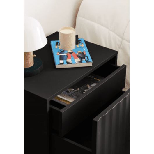 La mesita de noche Sierra 2C es una mesita de noche negra con un cajón abierto, que cuenta con una lámpara, una vela y un libro colorido en la parte superior. Diseñada por Cambres Design, ofrece un almacenamiento elegante, con una revista parcialmente visible escondida dentro del cajón.