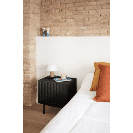 Un dormitorio minimalista destaca por una pared de ladrillos, la mesita de Noche Sierra 2C con una lámpara blanca y libros apilados. La cama luce un edredón blanco y almohadas de color naranja brillante. Un toque elegante de Cambres Design realza el espacio con piezas decorativas seleccionadas.