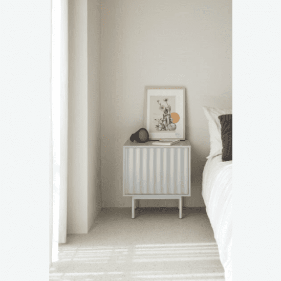 Una mesita de noche Sierra 2C con un patrón de rayas se encuentra contra la pared, adornada con obras de arte y un elegante objeto negro. Esta pieza de Cambres Design ofrece almacenamiento elegante, realzando el estilo de cualquier habitación, mientras que parte de una cama blanca también se ve cerca.