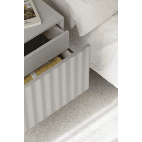 Una mesita de noche gris Sierra 2C de Cambres Design, con dos cajones abiertos para guardar libros con elegancia, se coloca junto a una cama con una manta acolchada blanca.