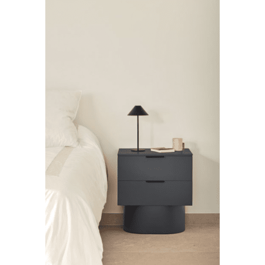 Un dormitorio moderno presenta una cama blanca a la izquierda y una elegante mesita de noche Totem 2C con dos cajones a la derecha. Este diseño contemporáneo incluye una pequeña lámpara negra, un libro y una taza beige, con un almacenamiento minimalista.