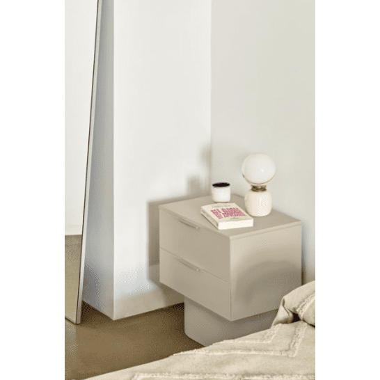 Un rincón minimalista del dormitorio muestra la mesita de noche Totem 2C con una lámpara, un libro y una vela. Un espejo reposa contra la pared junto a la cama adornada con una manta beige, que encapsula el diseño contemporáneo.