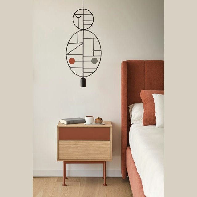 Un dormitorio moderno presenta una pared beige, arte abstracto y una cama tapizada en rojo. La mesita de noche Yoko 2C sostiene una taza, un objeto pequeño y libros, lo que agrega un toque de diseño contemporáneo.