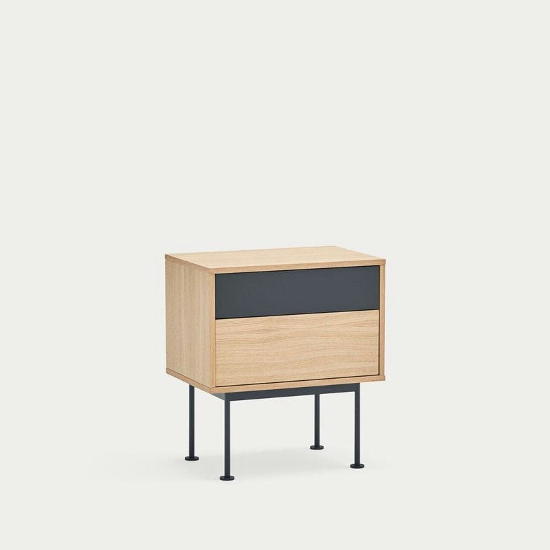 La mesita de noche Yoko 2C de Cambres Design es una mesita de noche contemporánea de madera con dos cajones, uno de ellos en un llamativo color negro. Tiene un acabado de madera clara y reposa sobre cuatro patas delgadas de metal negro sobre un fondo blanco liso.