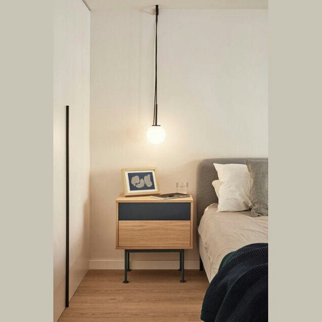 Un dormitorio moderno de Cambres Design cuenta con una mesita de noche Yoko 2C con un cajón azul, una pequeña foto enmarcada y un vaso. Una lámpara colgante ilumina el área junto a la cama adornada con ropa de cama gris y blanca, que personifica el diseño contemporáneo.