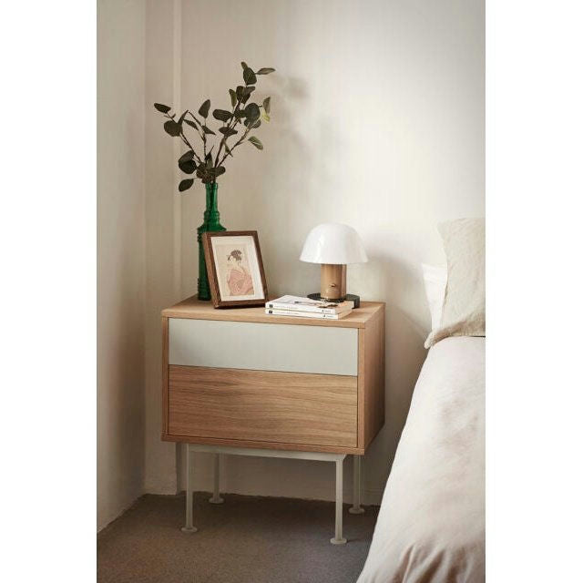 La mesita de noche Yoko 2C, con dos cajones, alberga con elegancia una planta verde embotellada, un cuadro enmarcado, libros y una lámpara blanca. Junto a una cama adornada con ropa de cama blanca, muestra el estilo contemporáneo de Cambres Design.