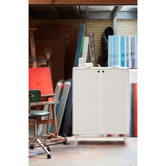 En un estudio de arte rodeado de cuadros y una silla, se encuentra el Aparador pequeño en metal con Puertas Punto 2P4C, sirviendo como mueble auxiliar.