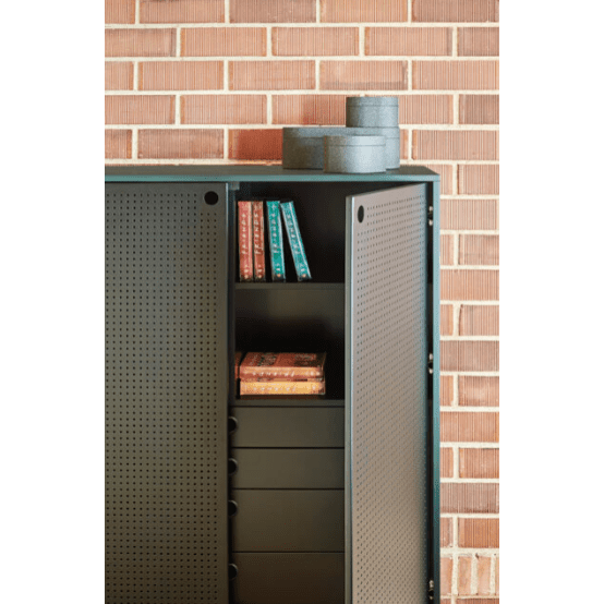 El Aparador pequeño de metal con Puertas Punto 2P4C, un mueble negro de diseño industrial, se apoya contra una pared de ladrillos. Presenta libros y cajas de almacenamiento en sus estantes, con cajas redondas elegantemente colocadas en la parte superior.