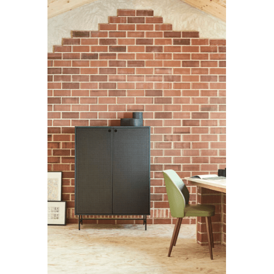 El Aparador pequeño de metal negro con puertas Punto 2P4C actúa como un elegante mueble auxiliar contra una pared de ladrillos, complementando perfectamente el escritorio de madera con su silla verde. Los cuadros enmarcados se apoyan sutilmente contra la pared en el suelo, realzando el estilo de diseño industrial.