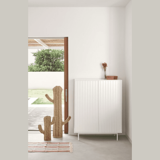 Una habitación minimalista presenta el Aparador pequeño en DM lacado Sierra 2P4C para almacenamiento práctico, dos elementos de decoración en forma de cactus y una alfombra de rayas cerca de una puerta corrediza abierta que revela una vista exterior con árboles y una pared blanca.