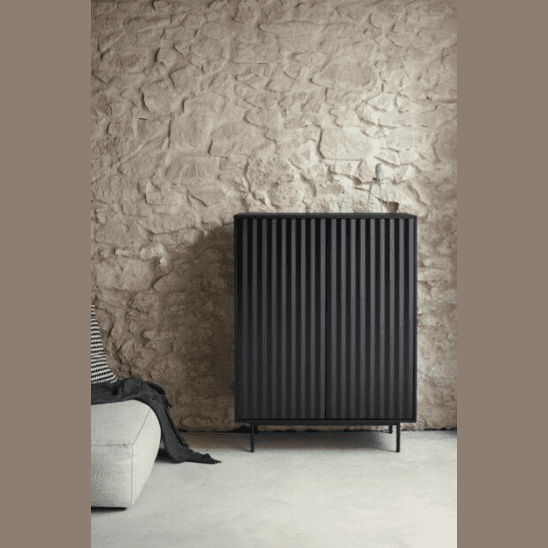 El Aparador pequeño en DM lacado Sierra 2P4C, en color negro con un diseño acanalado, se apoya contra una pared de piedra texturizada cerca de un sofá de color claro con un cojín de rayas, ofreciendo almacenamiento práctico para su espacio de estar.