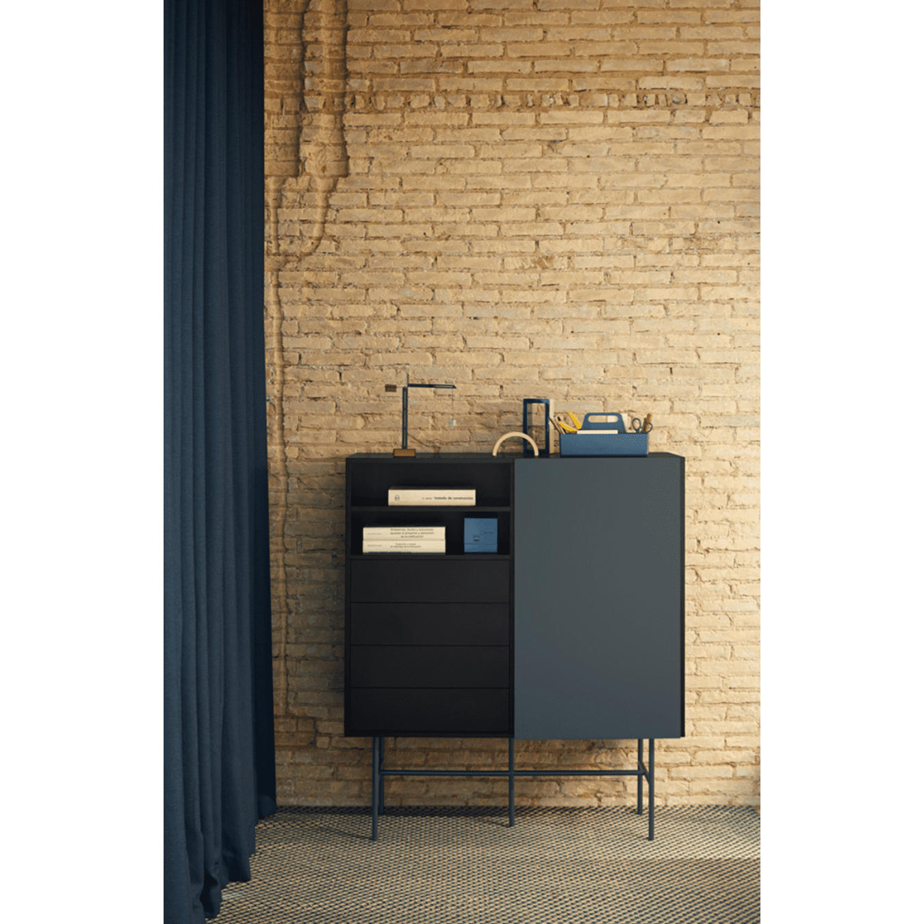 El Aparador con Puerta Corredera y Cajones Nube 1P4C, con estantes abiertos que exhiben libros y decoración, se encuentra contra una pared de ladrillos. Una cortina azul parcialmente visible a la izquierda le agrega encanto minimalista.