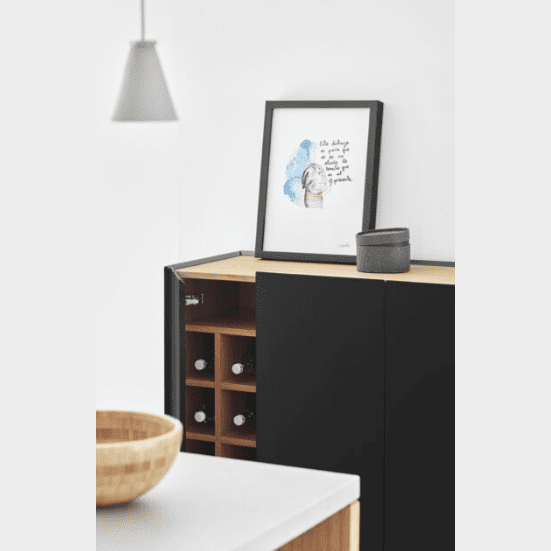 El Mueble Auxiliar con Puertas Arista 3P en elegante MDF lacado negro deja ver las botellas de vino a través de una puerta abierta. Un cuadro enmarcado y un pequeño recipiente gris añaden estilo en la parte superior, mientras que un cuenco de madera y una lámpara colgante completan la estética del diseño geométrico en primer plano.