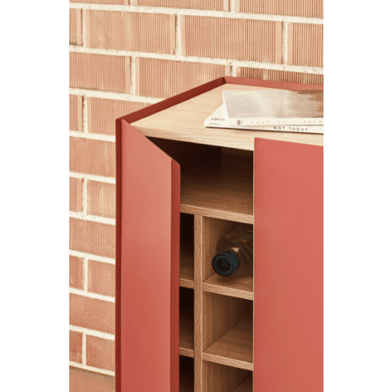 El Mueble Auxiliar con Puertas Arista 3P, colocado con estilo contra una pared de ladrillos, revela un espacio para guardar botellas de vino y una botella con las puertas abiertas. Un libro reposa sobre la parte superior, lo que resalta su diseño geométrico funcional y elegante.