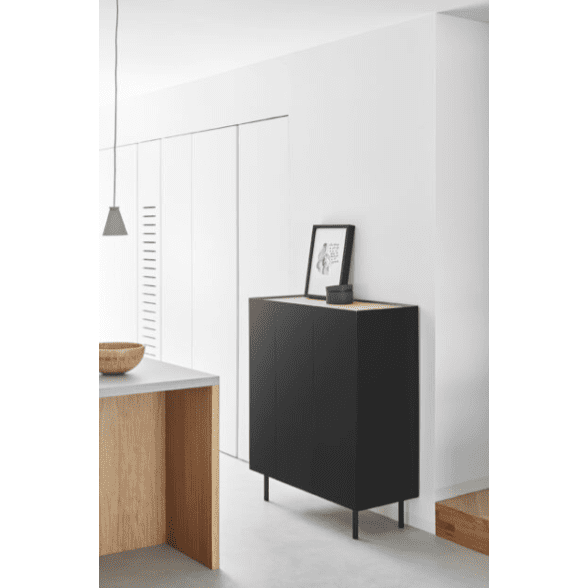 Un interior moderno y minimalista muestra el elegante Mueble Auxiliar con Puertas Arista 3P en color negro sobre patas delgadas, rematado con un cuadro enmarcado y un cuenco de madera. El estilo limpio se ve realzado por las paredes blancas y una lámpara colgante, mientras que el MDF lacado se suma al diseño elegante.