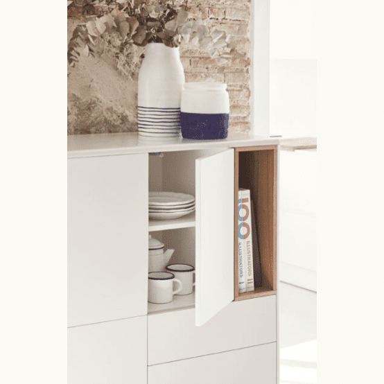 El gabinete Mueble Auxiliar con Puertas y Cajones Corvo 3P3C blanco tiene una puerta abierta con platos, tazas, un libro de cocina y frascos de cerámica con rayas azules cuidadosamente apilados en la parte superior. Ofrece una combinación perfecta de almacenamiento innovador y decoración contemporánea contra una pared de ladrillo rústico.