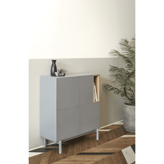 El Mueble Auxiliar con Puertas y Cajones Corvo 3P3C, un moderno mueble gris con almacenamiento innovador, exhibe libros en su estante. Ubicado sobre un piso de madera con formas geométricas, está adornado con un jarrón y una planta, junto a una planta alta en maceta contra una pared beige de dos tonos que muestra una decoración contemporánea.