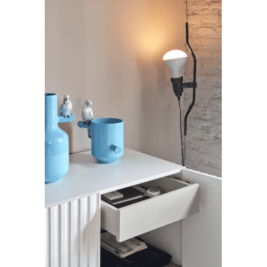 El Aparador pequeño con puertas Doric 2P2C es un mueble blanco elegante y moderno que funciona como un elegante mueble auxiliar y que exhibe un cajón abierto con jarrones azules y dos figuras de pájaros. Una bombilla encima está elegantemente montada en la pared de ladrillo.