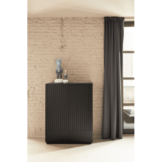 El Aparador pequeño con Puertas Doric 2P2C en color negro, con ranuras verticales, reposa sobre una pared beige texturizada. Dos pequeñas esculturas adornan su parte superior, lo que lo convierte en un elegante mueble auxiliar que complementa la gran ventana con cortinas oscuras a la derecha.