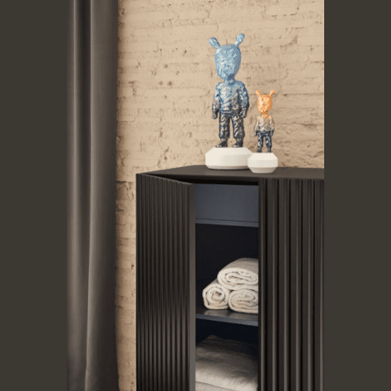 Dos esculturas abstractas con forma de conejo adornan un Aparador pequeño con Puertas Doric 2P2C con toallas dobladas. Una pared de ladrillos y una cortina sirven de fondo, creando una sorprendente combinación de diseño contemporáneo.