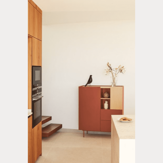 Una cocina minimalista con gabinetes de madera, hornos empotrados y un pequeño aparador Aparador pequeño con Puertas Otto 2P2C adornado con una figura de pájaro negro y jarrones de cerámica; una encimera beige realza esta elegancia moderna de Cambres Design.