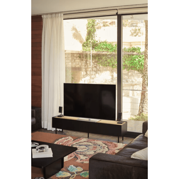 La sala de estar cuenta con un televisor de pantalla plana grande en el Mueble TV Arista 1P2C con almacenamiento elegante, acompañado de parlantes. Una alfombra colorida le agrega vitalidad al piso, mientras que la luz natural se cuela a través de una gran ventana con cortinas transparentes, mostrando un diseño moderno.