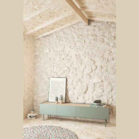 En una habitación con paredes de piedra y un techo inclinado de madera, el Mueble TV Arista 1P2C actúa como un sofisticado espacio de almacenamiento. Dos jarrones y obras de arte enmarcadas descansan elegantemente sobre él. Una alfombra colorida es parcialmente visible en el piso, completando el diseño contemporáneo de este acogedor espacio.