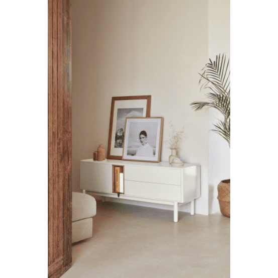 El Mueble TV Corvo 1P2C, colocado contra una pared de color crema, ofrece almacenamiento funcional con fotografías enmarcadas y decoración. A la derecha hay una planta en maceta junto a una puerta de madera, lo que crea un diseño contemporáneo que complementa el espacio a la perfección.