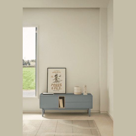 En una habitación minimalista con paredes de color beige claro, un Mueble TV Corvo 1P2C en azul ofrece espacio de almacenamiento funcional junto con detalles decorativos debajo de un cuadro. Una gran ventana a la izquierda revela un paisaje verde en el exterior, que personifica el diseño contemporáneo.