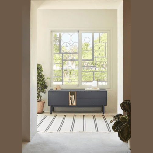 Una habitación moderna cuenta con una consola de TV Corvo 1P2C gris de Mueble TV que ofrece espacio de almacenamiento funcional y una alfombra de rayas. Dos plantas en macetas aportan frescura, mientras que una gran ventana geométrica ofrece vistas de los árboles, lo que realza el diseño contemporáneo.