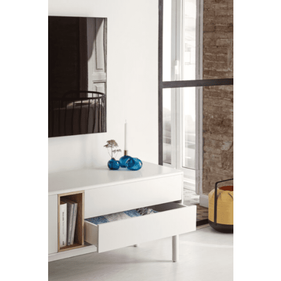 El Mueble TV Corvo 2P2C blanco ofrece almacenamiento versátil con un cajón abierto que exhibe libros, una vela, una decoración de vidrio azul y una planta en maceta. Un televisor montado en la pared está enmarcado por una ventana y una pared de ladrillos, lo que completa el diseño contemporáneo sin fisuras.