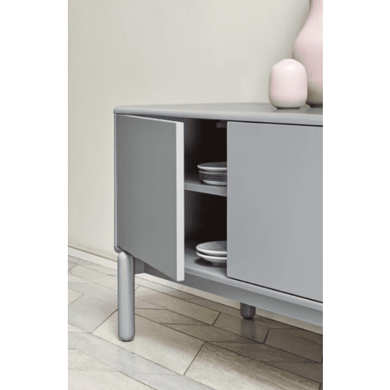 El Mueble de TV Corvo 2P2C, un moderno mueble gris con una puerta entreabierta y platos apilados en su interior, se encuentra sobre un piso de madera clara. Adoptando el diseño contemporáneo, se complementa con dos jarrones rosas en la parte superior.