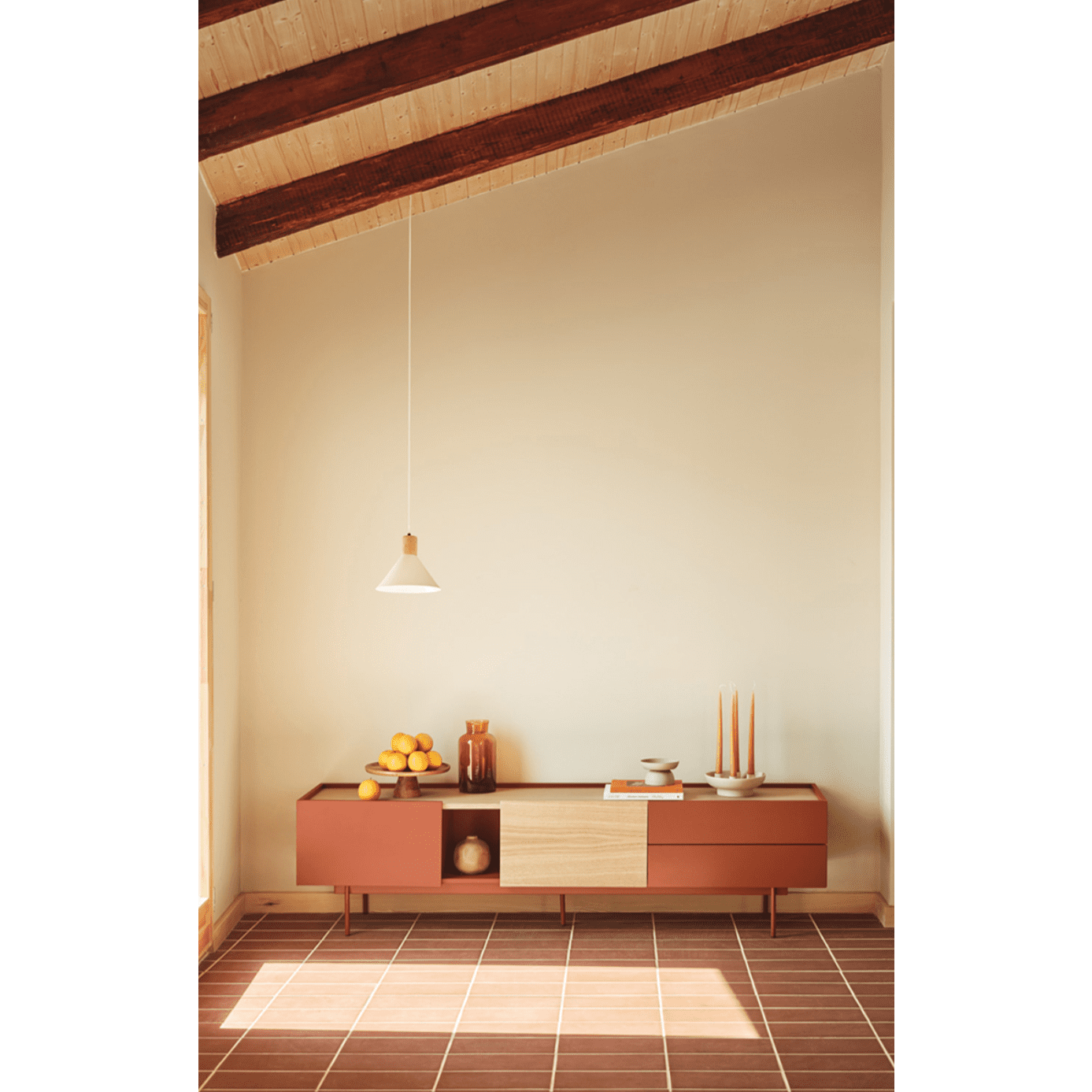 Una habitación minimalista cuenta con el Mueble TV Otto 2P2C para almacenar multimedia, cuencos de fruta, velas y una lámpara colgante. El espacio combina el encanto rústico con el diseño contemporáneo a través de vigas de madera a la vista y un piso de baldosas.