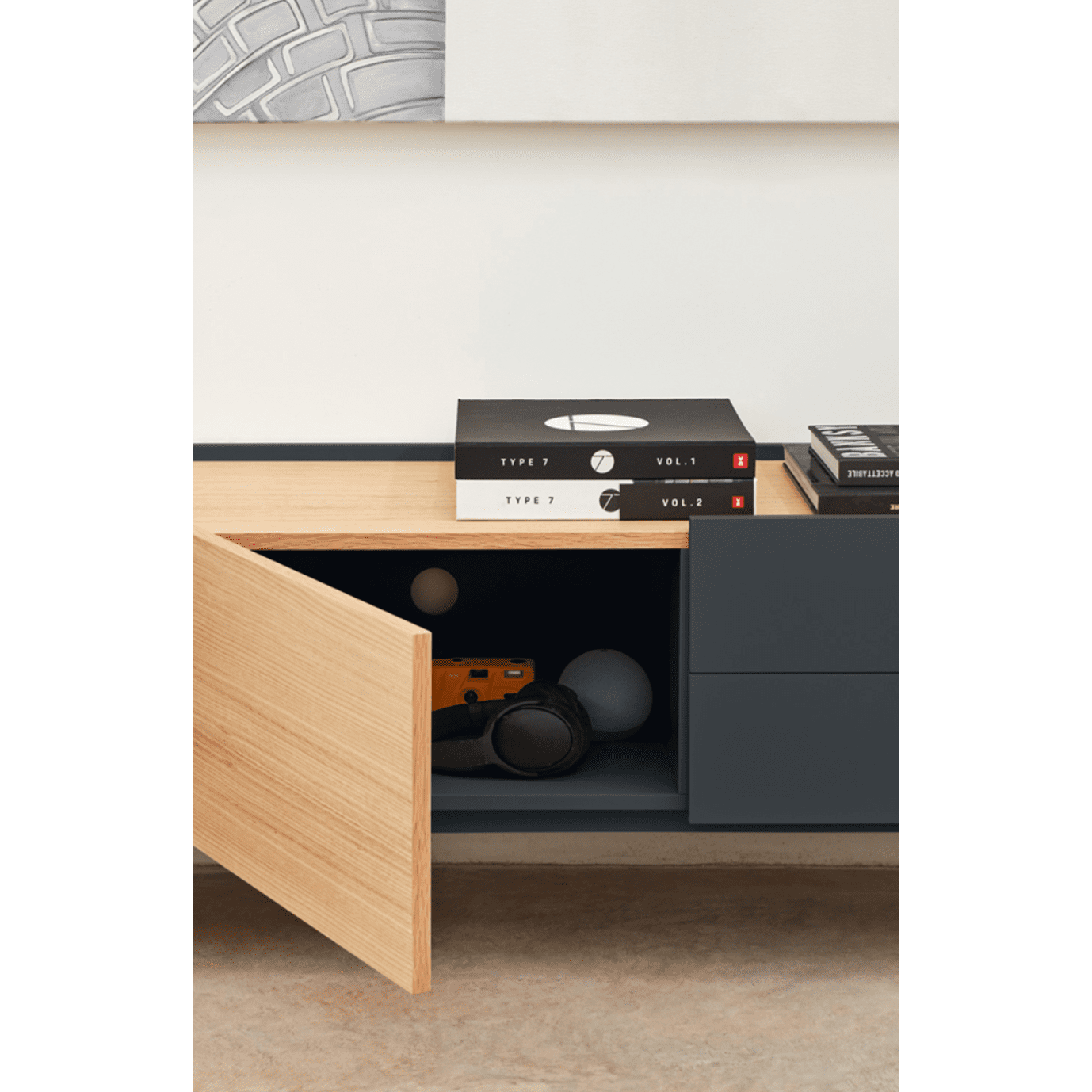 El Mueble TV Otto 2P2C, de diseño minimalista, cuenta con un mueble de madera con una puerta abierta que revela una cámara, una bola blanca y un objeto naranja en su interior. Dos libros y una revista están perfectamente apilados en la parte superior, lo que lo hace ideal para almacenar multimedia.