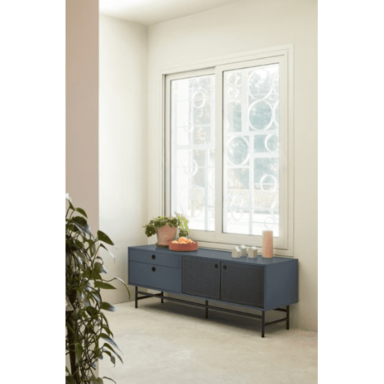 El Mueble TV Punto 2P2C, un elegante aparador azul con cuatro puertas y un cajón, se encuentra junto a una gran ventana en una habitación luminosa. El diseño moderno se combina con un elegante espacio de almacenamiento, ya que los elementos decorativos y una planta en maceta lo complementan, mientras que una planta frondosa aporta vitalidad a la escena.