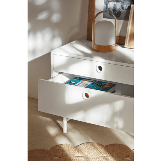 El Mueble TV Punto 2P2C, que ofrece un almacenamiento elegante, incluye dos cajones con tiradores circulares. El cajón inferior está abierto y revela un libro azul. Una linterna y un cuadro enmarcado se encuentran en la parte superior bajo la luz del sol, lo que resalta su diseño moderno.