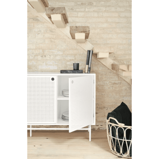 Un interior moderno muestra el Mueble TV Punto 2P4C, un gabinete perforado blanco debajo de una escalera de madera, que ejemplifica el diseño contemporáneo. Las puertas abiertas revelan platos organizados para almacenamiento y organización. Una canasta negra y libros cercanos realzan el ambiente elegante.