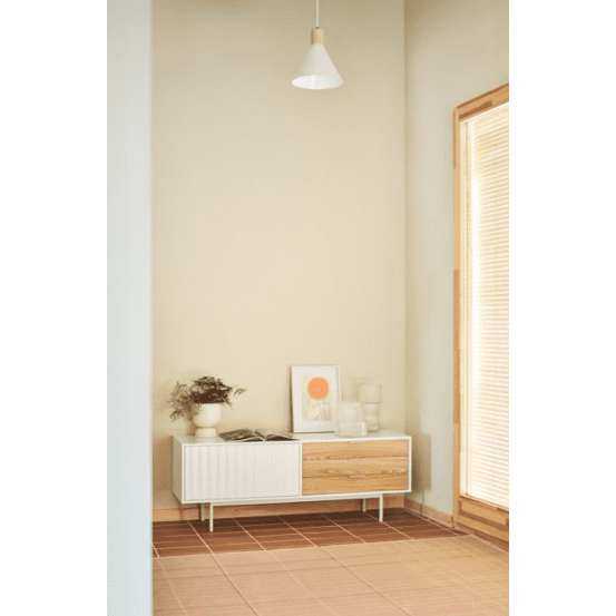 En un rincón minimalista se exhibe el Mueble TV Sierra 1P3C junto a un cuadro, una planta exuberante y una lámpara colgante. Las baldosas del piso iluminadas por el sol resaltan el diseño asimétrico con ventanas elegantemente cubiertas por persianas.