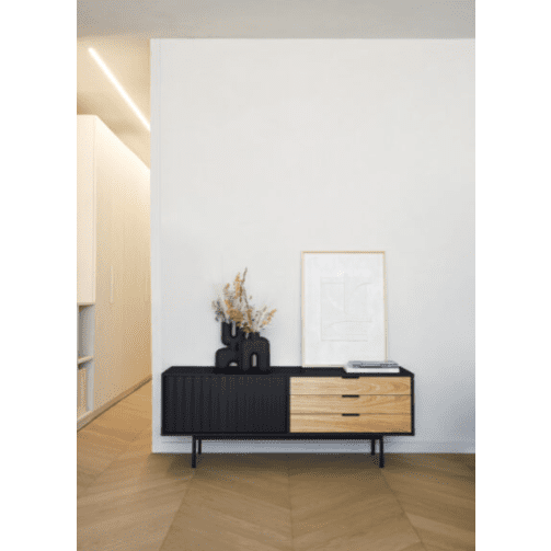El Mueble TV Sierra 1P3C, que muestra su moderno diseño en negro y madera con diseño asimétrico, se apoya contra una pared blanca adornada con un arreglo de plantas secas y una obra de arte minimalista enmarcada en la parte superior.