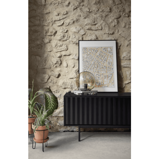 Una habitación minimalista cuenta con una pared de piedra texturizada, un elegante aparador negro que también funciona como Mueble TV Sierra 2P3C, ilustraciones de mapas enmarcadas, plantas en macetas y una elegante esfera decorativa que se combinan a la perfección con el diseño moderno.