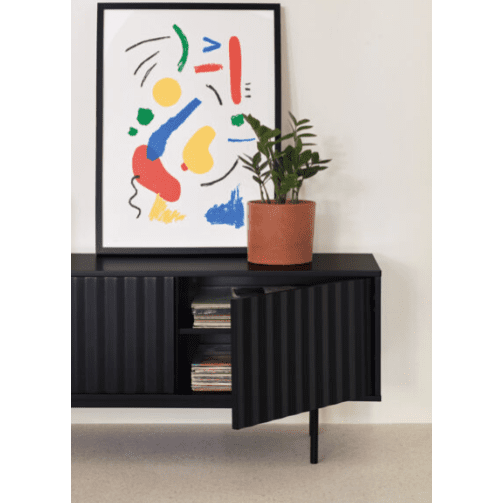 Un cuadro abstracto colorido se apoya en una pared sobre un aparador negro, resaltando su elegante diseño modernista. El Mueble TV Sierra 3P cuenta con una puerta abierta para un almacenamiento multimedia perfecto, complementada con una planta en maceta que le agrega encanto natural en la parte superior.