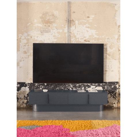Un televisor de pantalla plana montado en una pared envejecida y texturizada sobre un elegante Mueble TV Totem 4P con un acabado moderno y oscuro se complementa con una alfombra colorida en el piso de concreto.