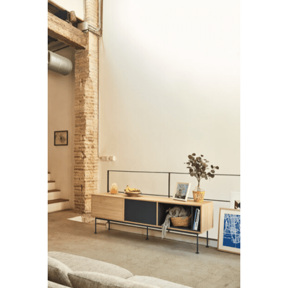 En una sala de estar contemporánea, el Mueble TV Yoko 1P2C reposa contra una pared blanca, adornado con elementos decorativos y marcos de fotos inclinados con estilo. Esta pieza ofrece un almacenamiento elegante y de buen gusto, realzando el diseño contemporáneo. Se ve una escalera a la izquierda.