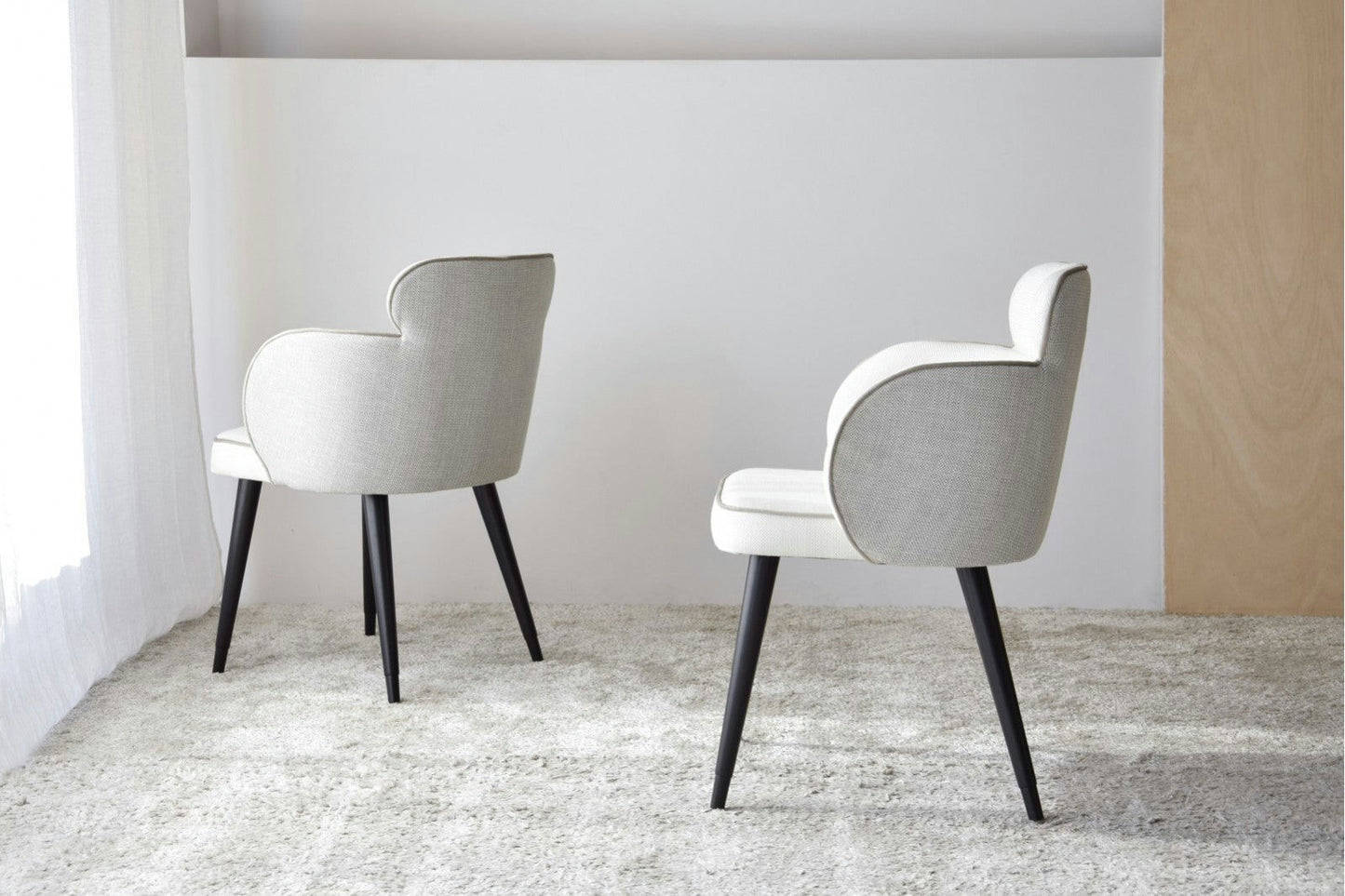 El Pack 2 Sillas de Comedor Tapizadas con Reposabrazos Oslo, con respaldos curvos y patas negras, está colocado sobre una alfombra texturizada contra una pared blanca con un panel de madera, mostrando un diseño escandinavo.