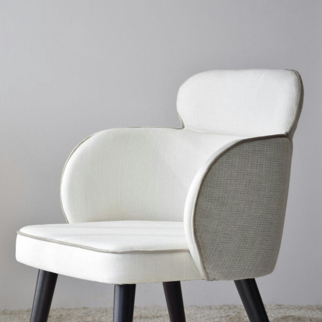 El Pack 2 Sillas de Comedor Tapizadas con Reposabrazos Oslo presenta una tapicería blanca moderna, un respaldo curvo y patas de madera oscura, que personifican el diseño escandinavo. Estas sillas descansan sobre una alfombra clara contra una pared lisa.