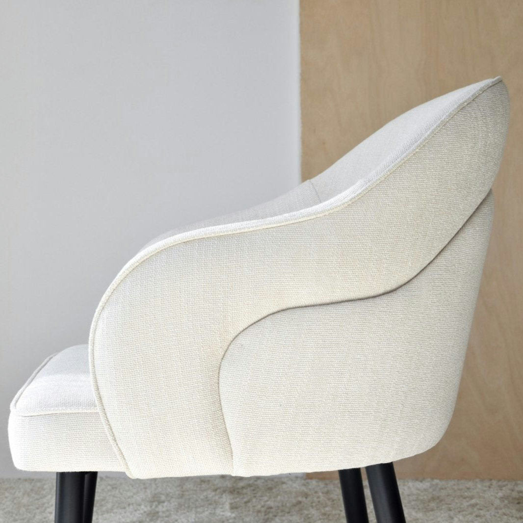 El Pack 2 Sillas de Comedor con Reposabrazos Roma (Beige claro) muestra un diseño ergonómico moderno con su respaldo curvo tapizado y patas negras, descansando elegantemente sobre una alfombra de textura ligera.